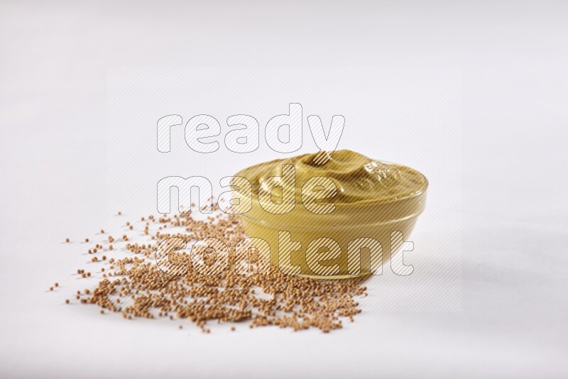 A glass bowl full of mustard paste with mustard seeds underneath on white flooring