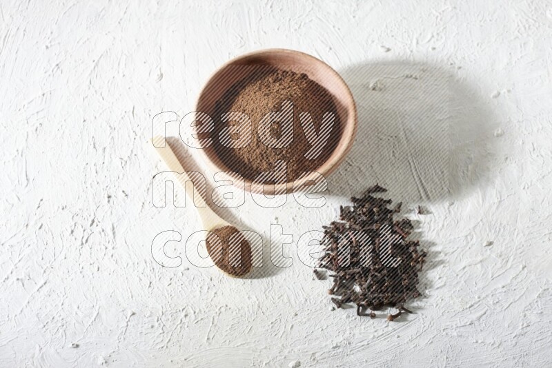 A wooden bowl and wooden spoon full of cloves powder with cloves spread on textured white flooring