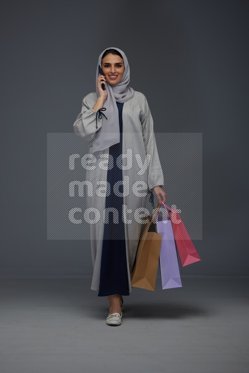 A Saudi woman wearing a light gray Abaya and head scarf standing and holding shopping bags making different poses eye level on a grey background