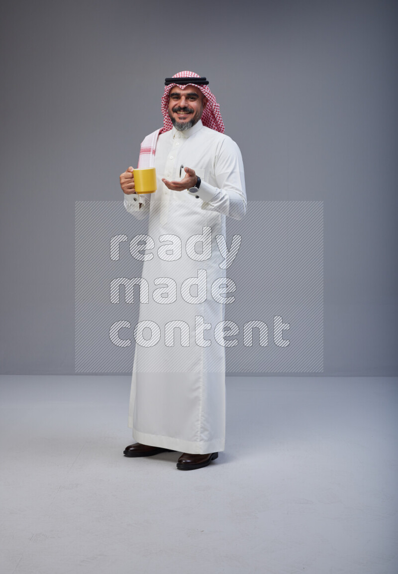 Saudi man Wearing Thob and red Shomag standing holding mug on Gray background