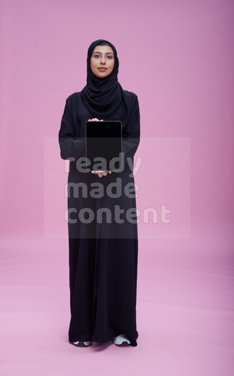 Saudi woman wearing Abaya standing showing tablet to camera on pink background