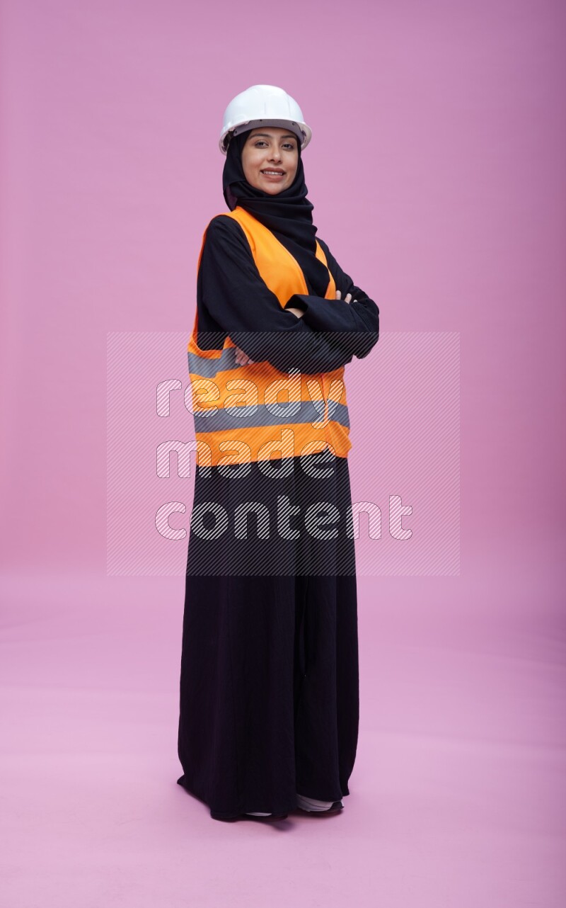 Saudi woman wearing Abaya with engineer vest and helmet standing with crossed arms on pink background