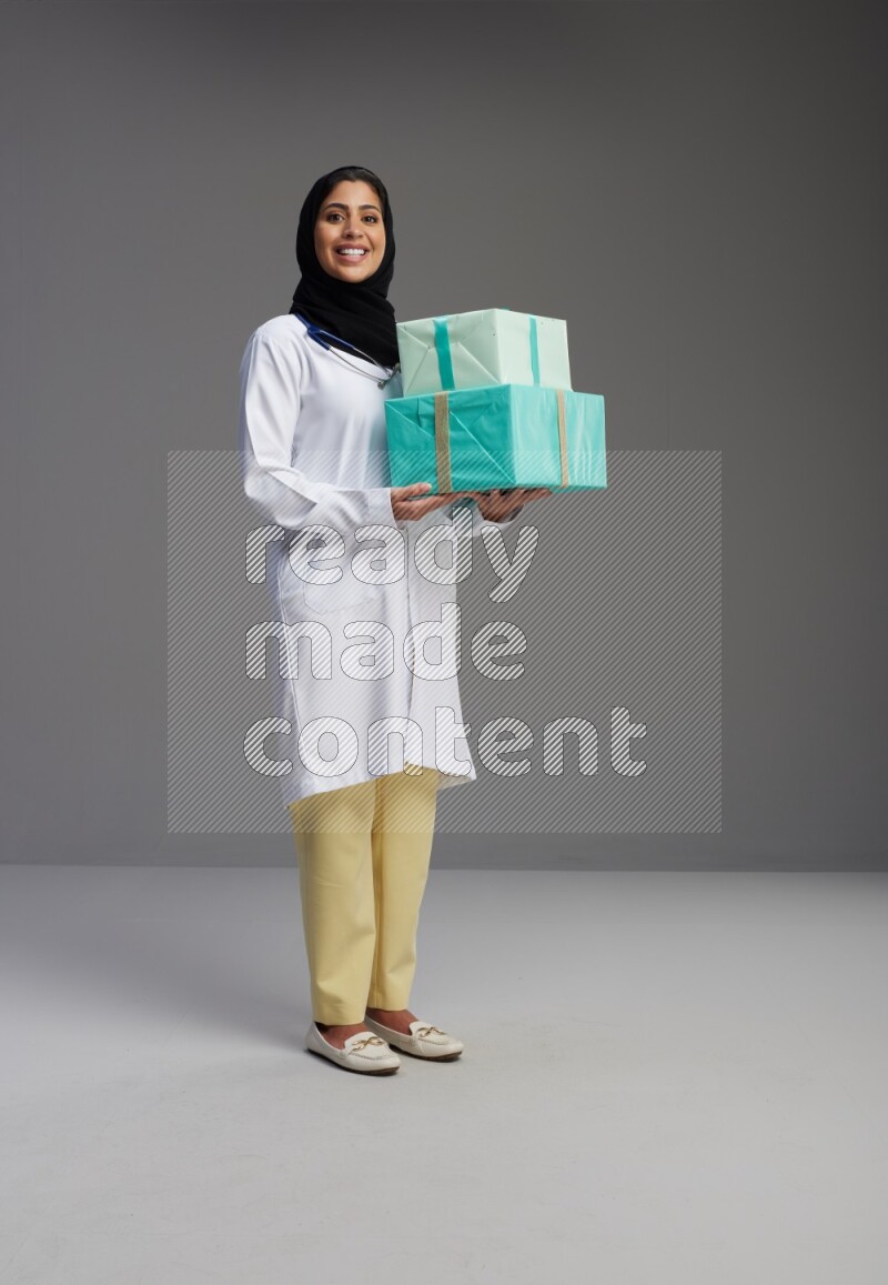 Saudi woman wearing lab coat with stethoscope standing holding gift box on Gray background