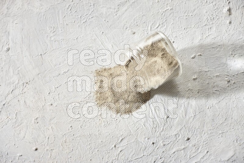 A flipped glass jar full of white pepper powder with spilled powder on textured white flooring