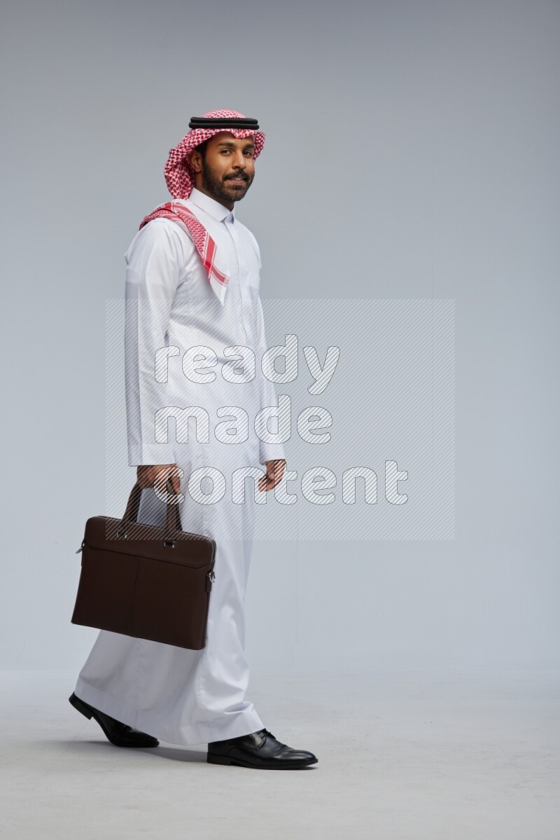 Saudi man Wearing Thob and shomag standing holding bag on Gray background