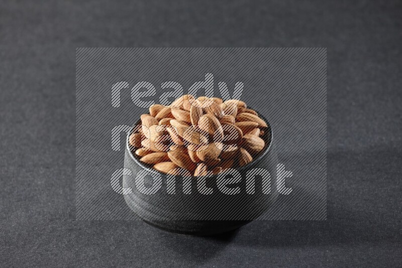 A black pottery bowl full of peeled almonds on a black background in different angles