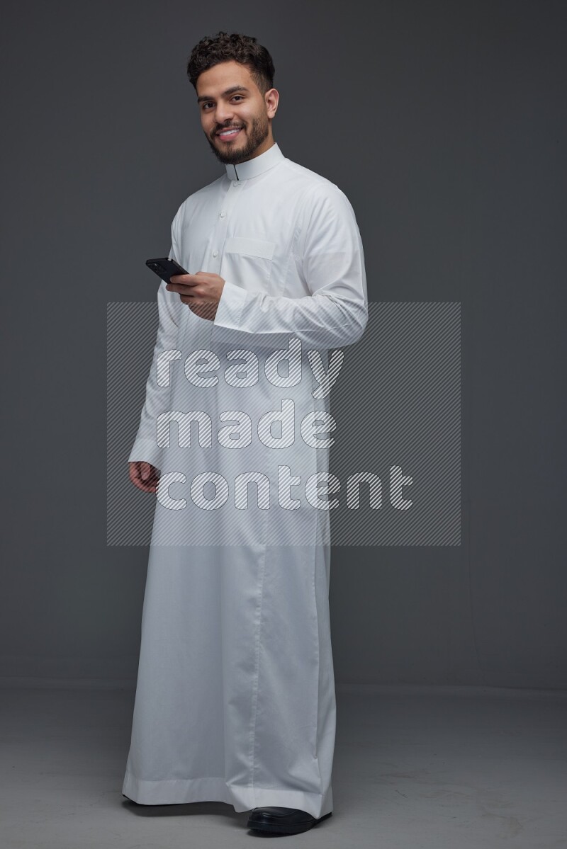 A Saudi man wearing Thobe standing and using his phone eye level on a gray background