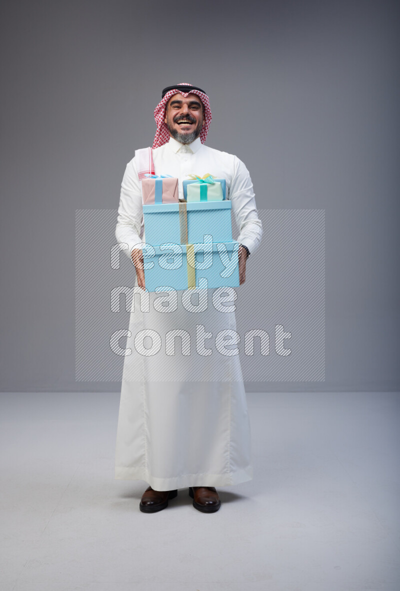 Saudi man Wearing Thob and red Shomag standing holding gift box on Gray background