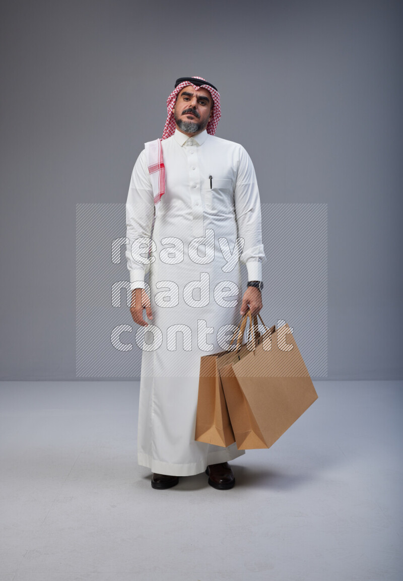 Saudi man Wearing Thob and red Shomag standing holding shopping bag on Gray background