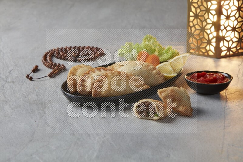 Four fried sambosas in an oval shaped black plate, beside a cut cheese sambosa, a brown misbaha and a golden lantern on a gray background