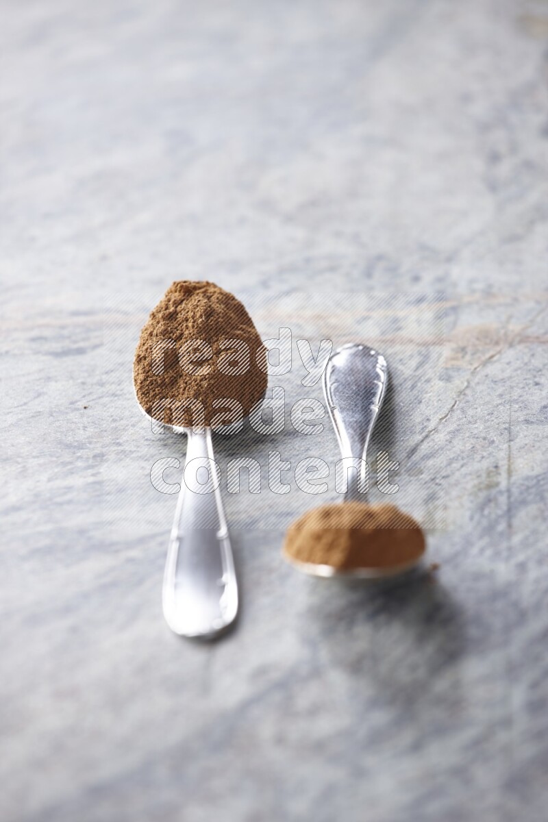 Two metal spoons full of cinnamon powder on grey marble background