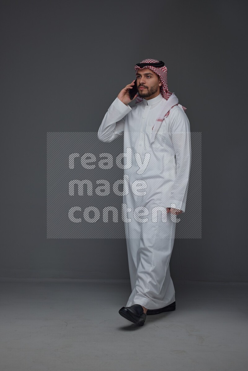 A Saudi man wearing Thobe and Shmagh talking in the phone eye level on a gray background