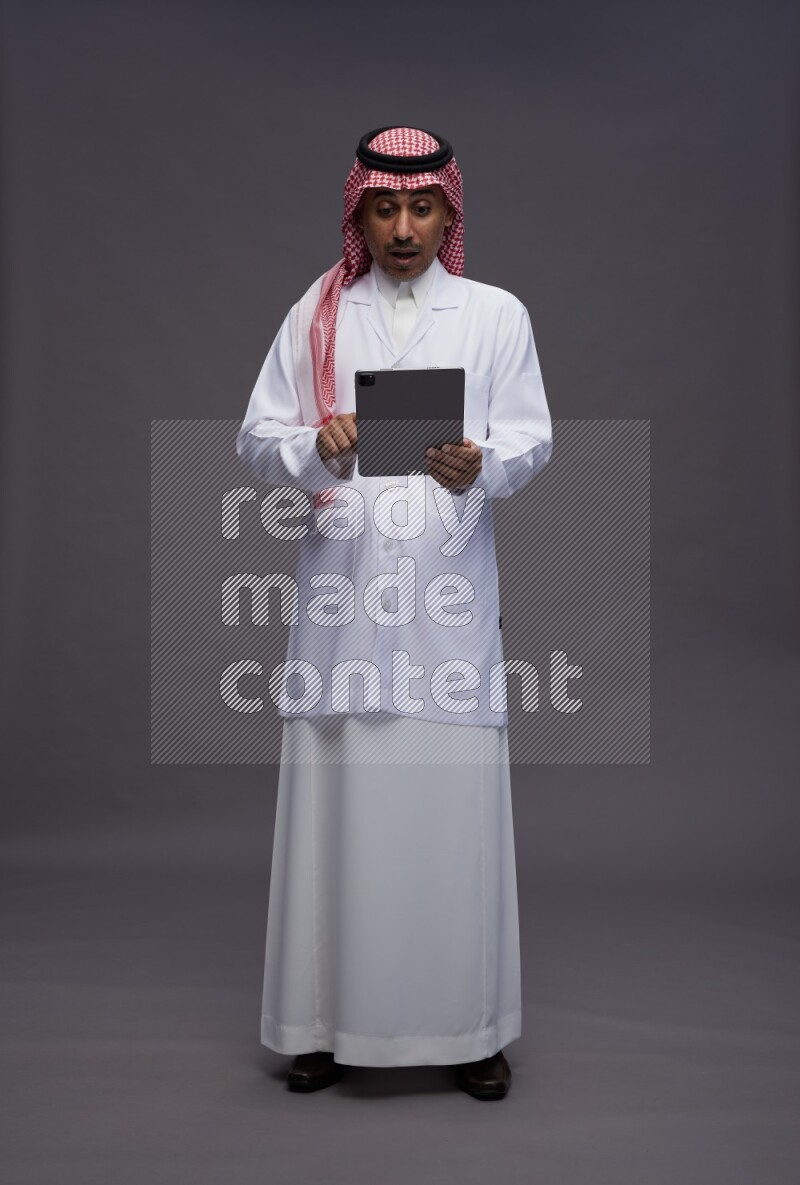 Saudi man wearing thob with lab coat and shomag with pocket employee badge standing working on tablet on gray background