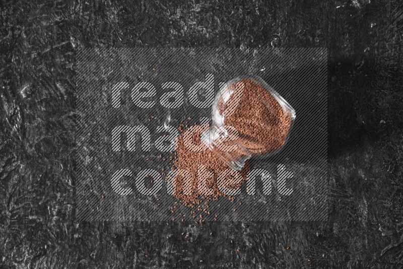 A glass spice jar full of garden cress seeds flipped and seeds spread out a textured black flooring