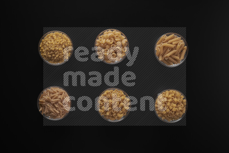 Different pasta types in 6 glass bowls on black background