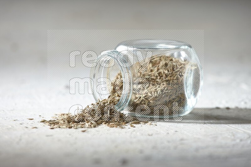 A flipped glass spice jar full of cumin seeds and the seeds spilled out on textured white flooring