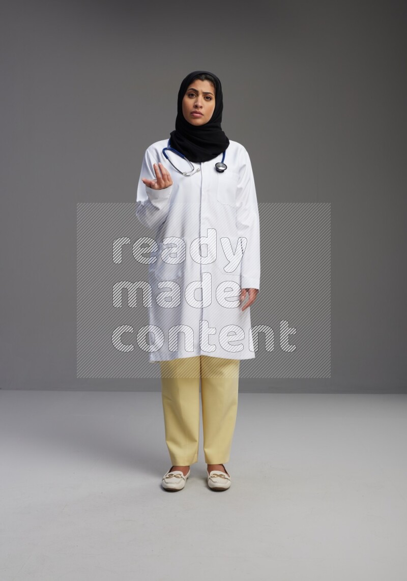 Saudi woman wearing lab coat with stethoscope standing interacting with the camera on Gray background