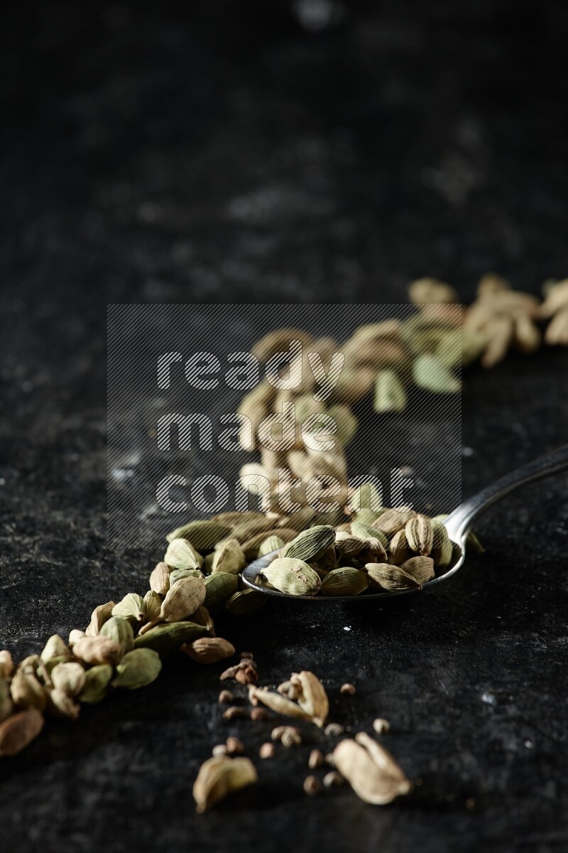A Metal spoon full of cardamom seeds and some seeds beside it on a textured black flooring