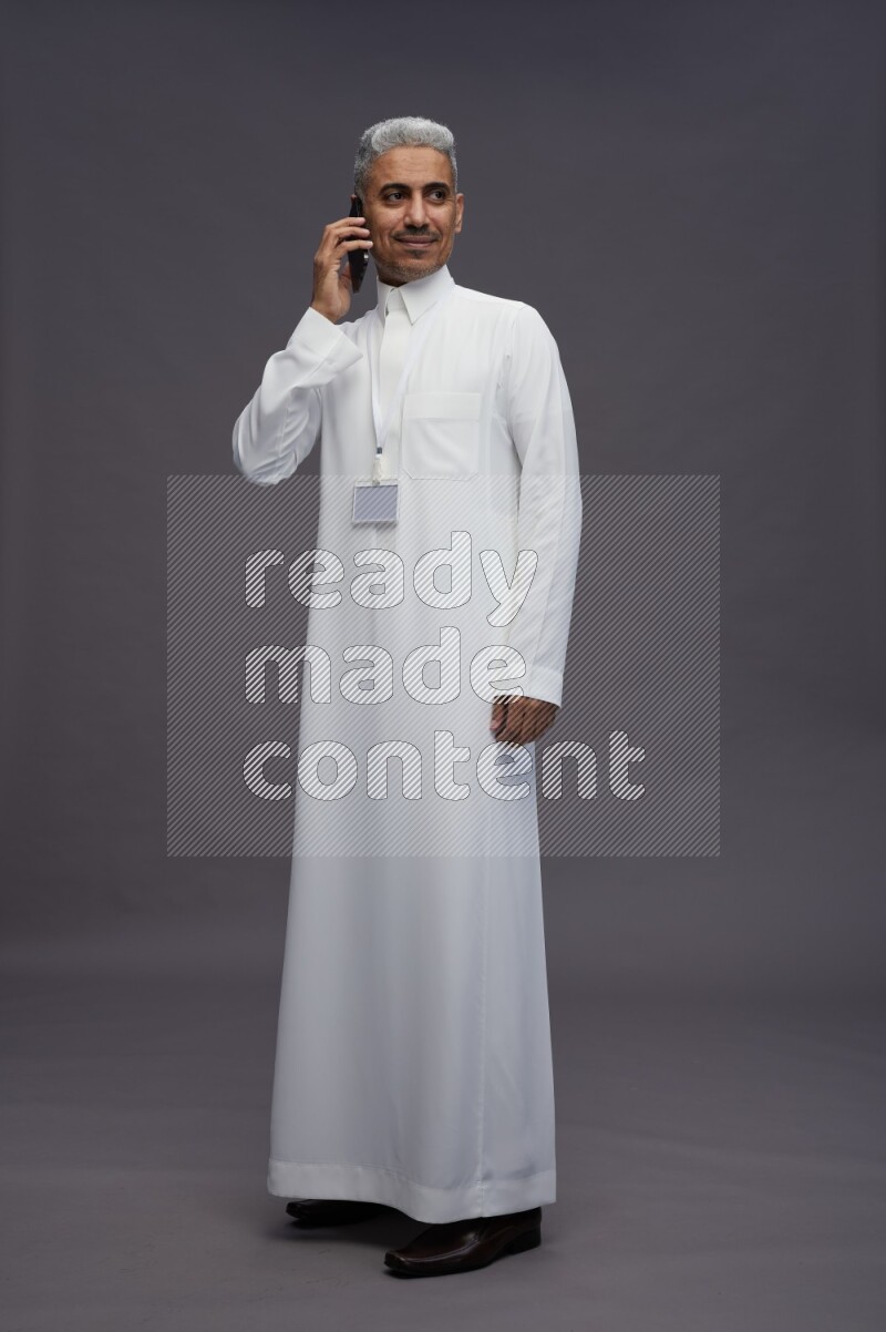 Saudi man wearing thob with neck strap employee badge standing talking on phone on gray background