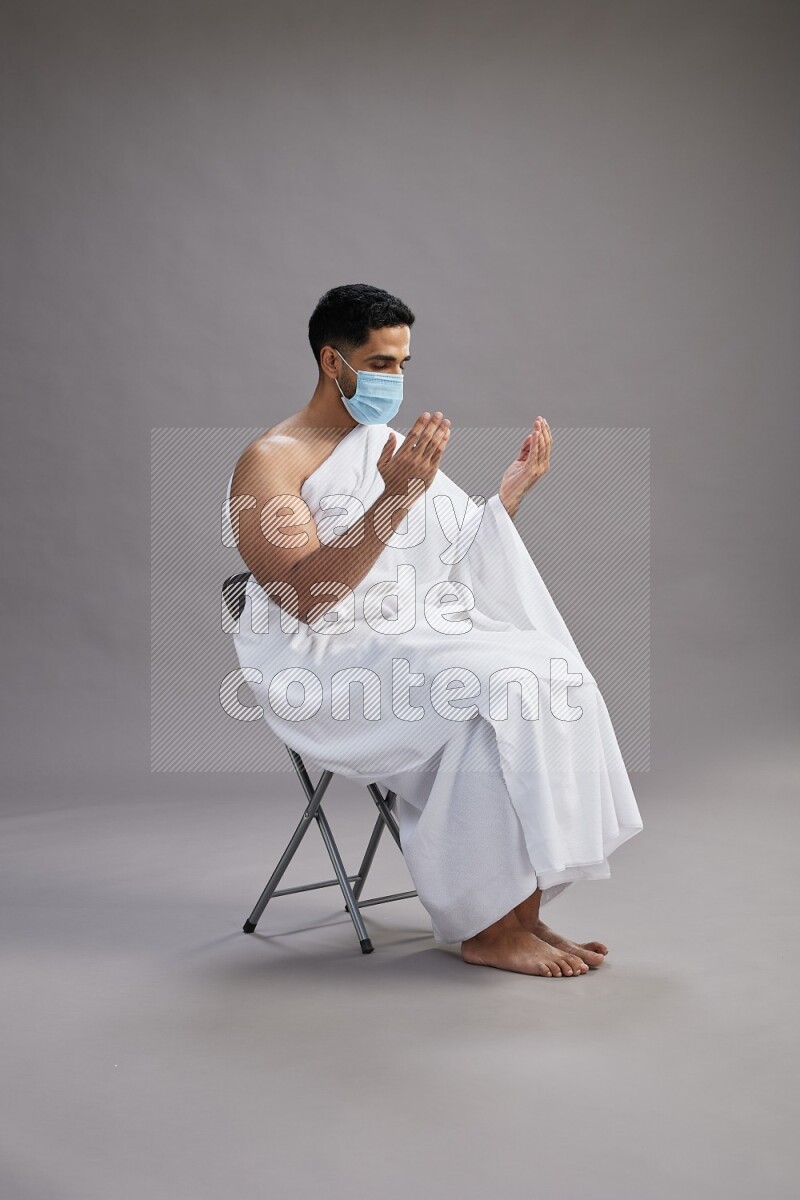 A man wearing Ehram with face mask sitting on chair dua'a on gray background