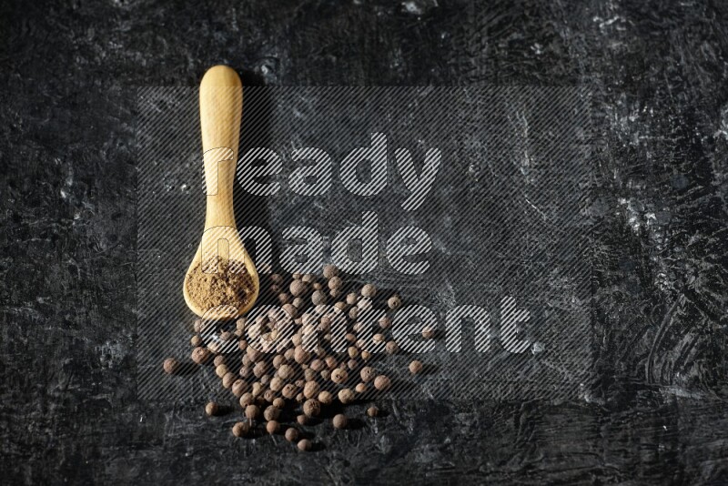 A wooden spoon full of allspice powder and whole balls spreaded on a textured black flooring