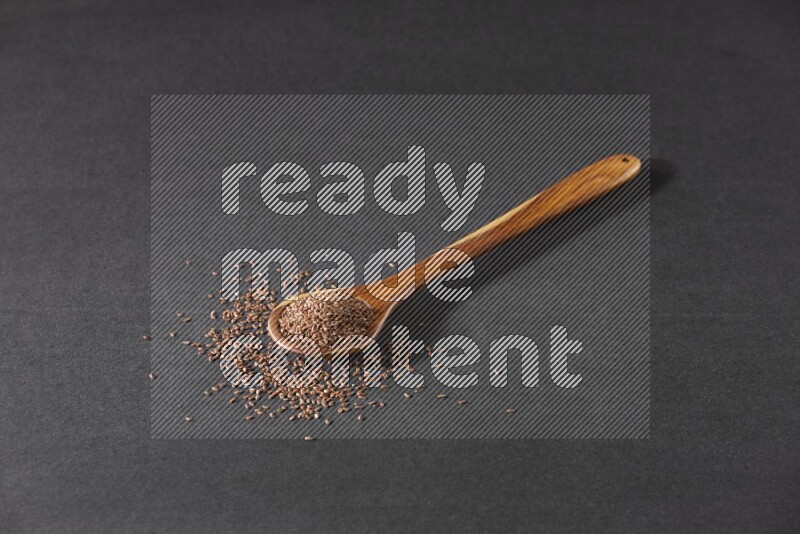 A wooden ladle full of flaxseeds and seeds spread beside it on a black flooring