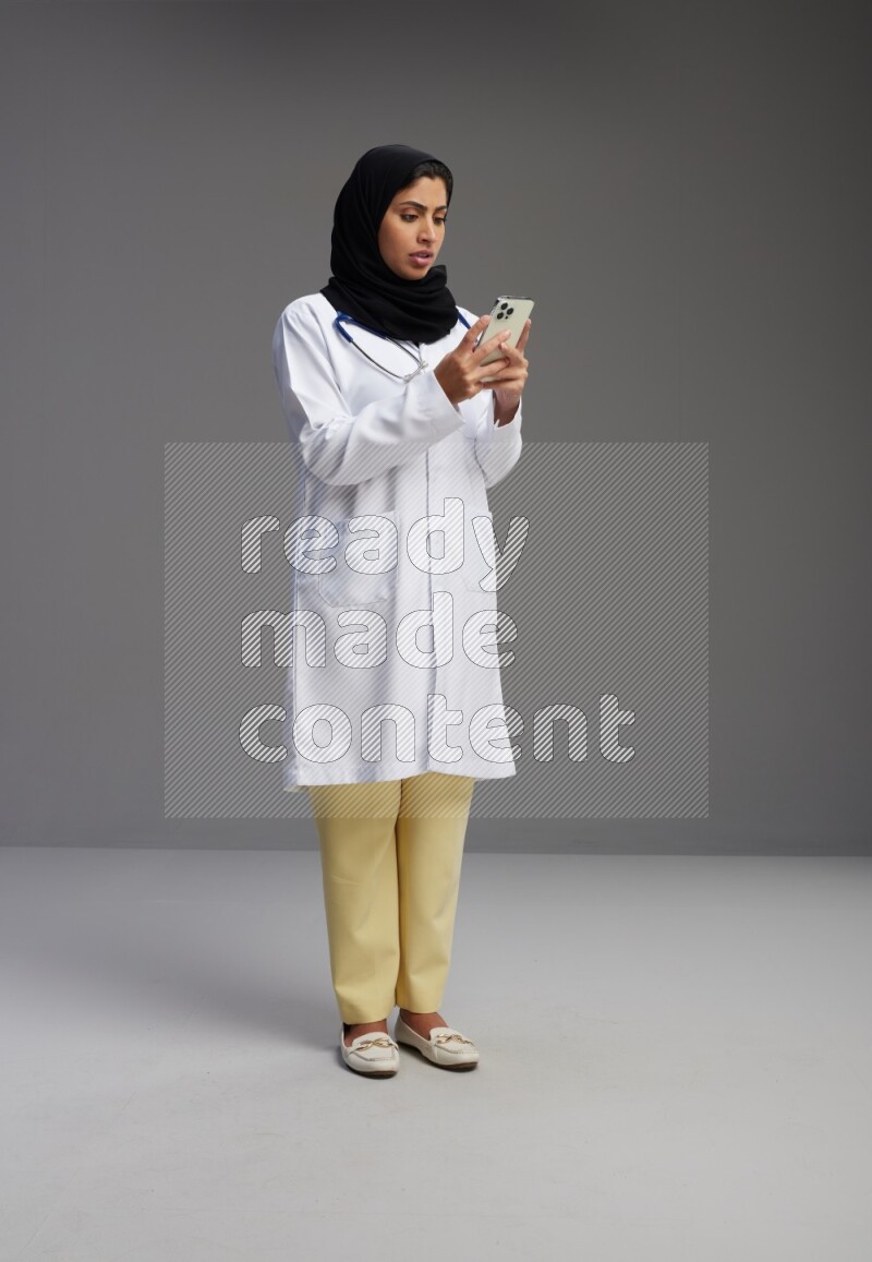 Saudi woman wearing lab coat with stethoscope standing texting on phone on Gray background