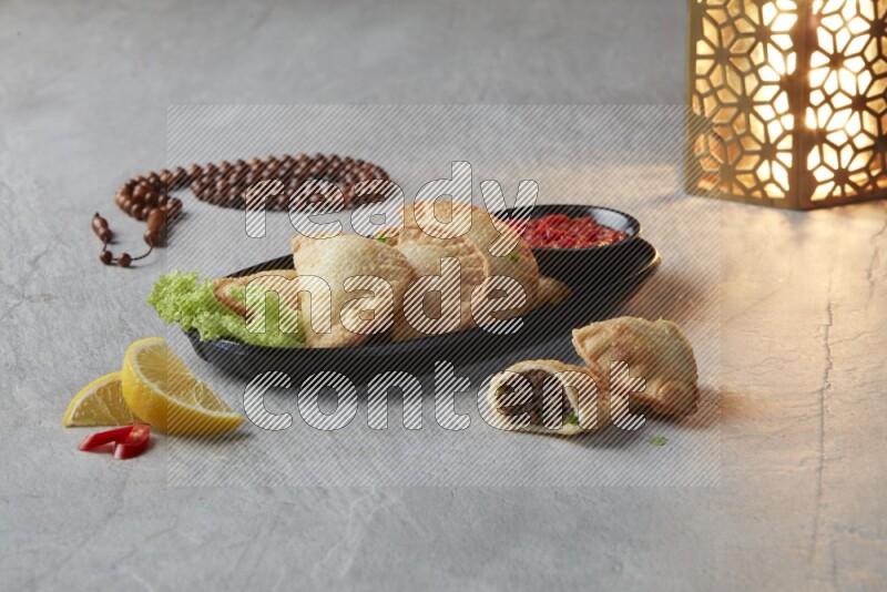 Five fried sambosas in an oval shaped black plate, beside a cut meat sambosa, a brown misbaha and a golden lantern on a gray background
