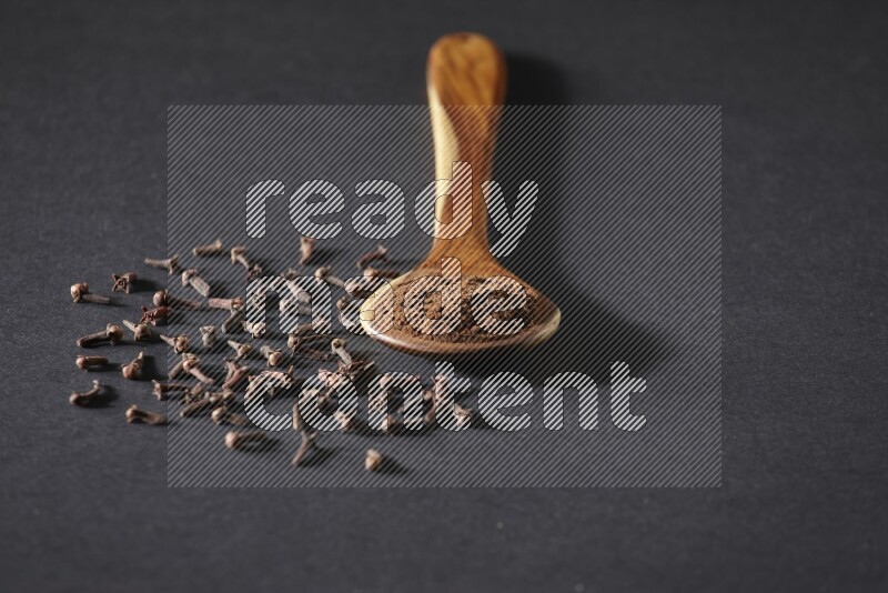 A wooden ladle full of cloves powder and some of whole cloves around it on a black flooring