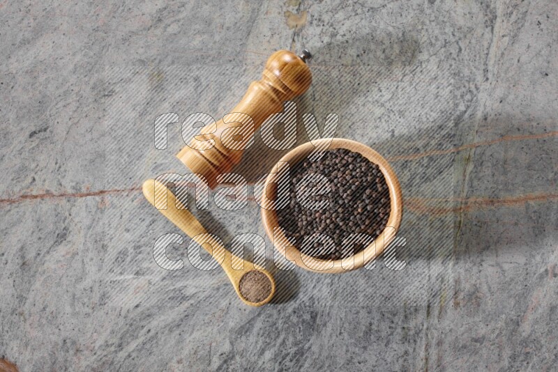 A wooden bowl full of black pepper and a wooden spoon full of black pepper powder and a wooden grinder on a marble flooring