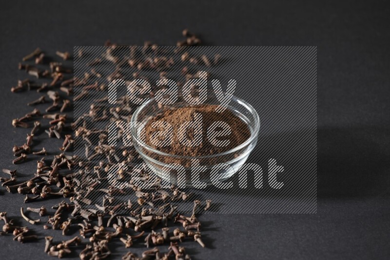 A glass bowl full of cloves powder with cloves grains spread on black flooring