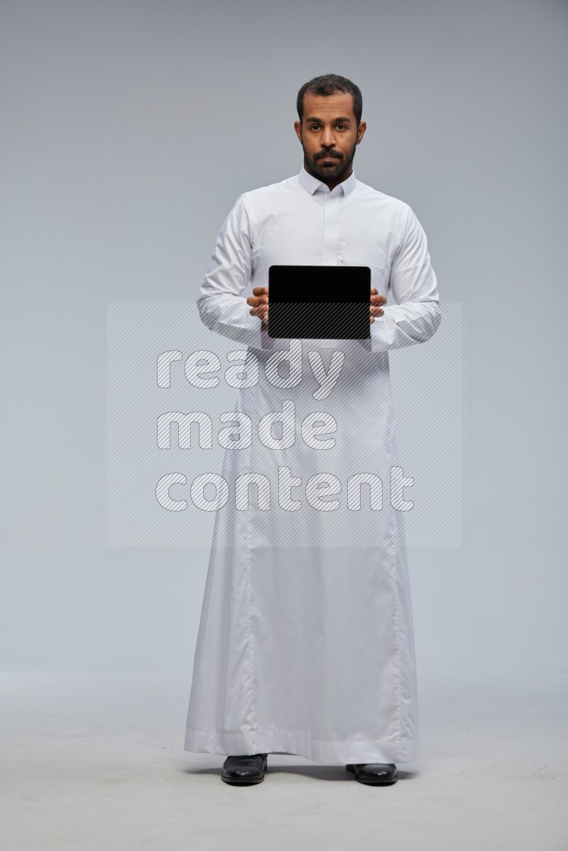 Saudi man wearing Thob standing showing tablet to camera on Gray background
