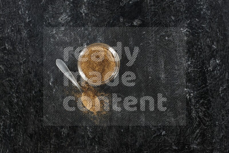 A glass jar and a metal spoon full of cumin powder on a textured black flooring