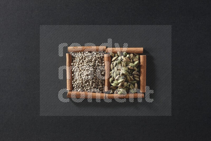 مربعان من أعواد القرفة ممتلئان بحبوب الفلفل الأبيض وحبوب الهيل علي خلفية سوداء