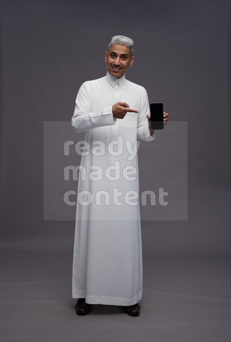 Saudi man wearing thob standing showing phone to camera on gray background