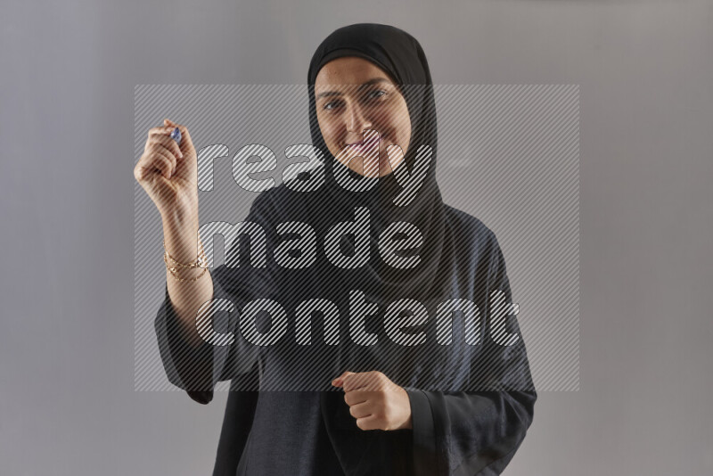 Woman in black abaya and hijab holding a marker pen to write on a transparent board with different reactions and expressions on grey background