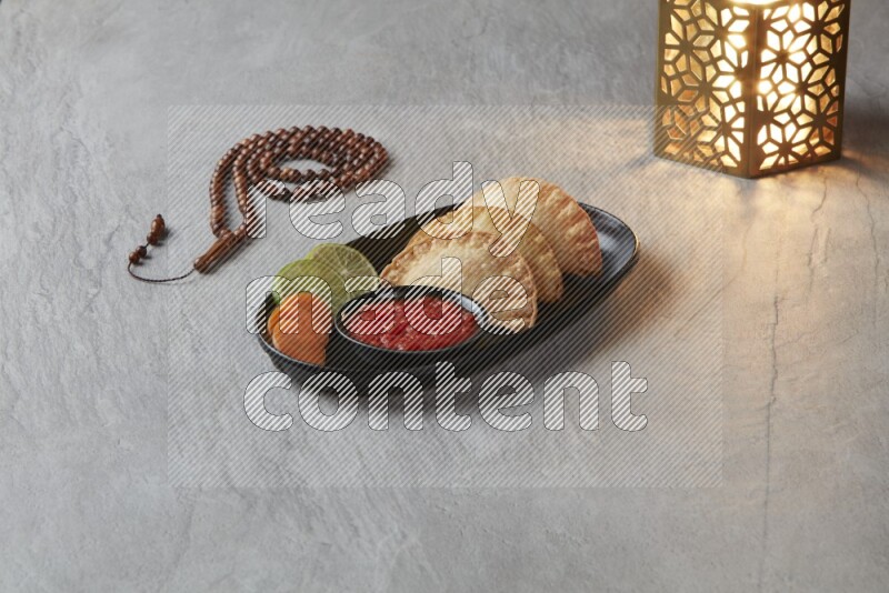 Three fried sambosas in an oval shaped black plate and a red sauce in a black round ramekin with a brown misbaha and a golden lantern on the side on a gray background