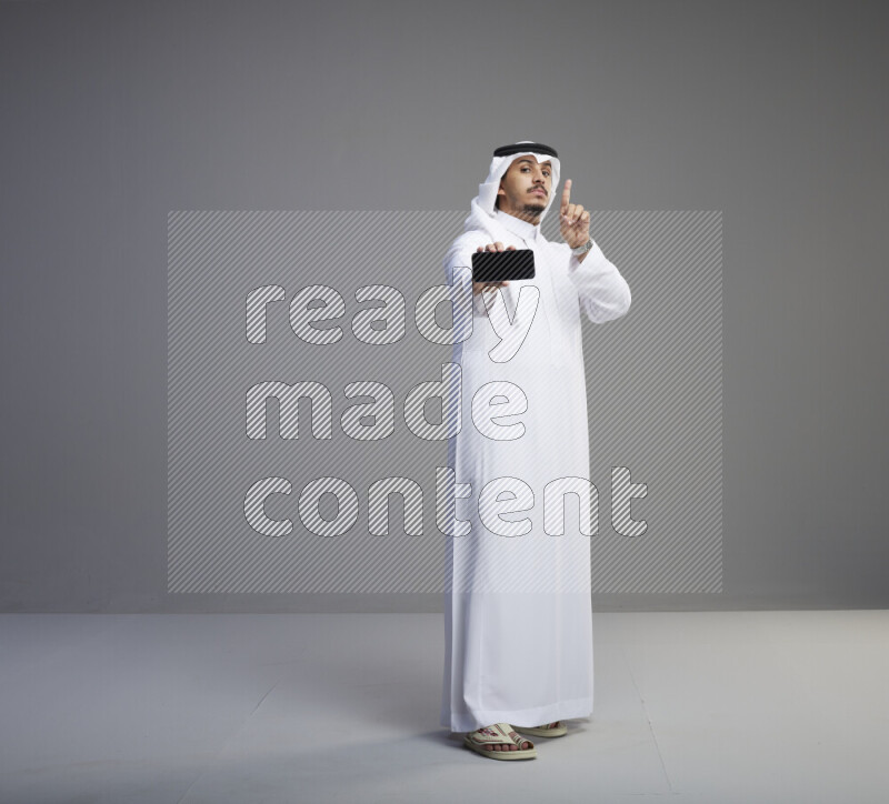 A Saudi man standing wearing thob and white shomag showing phone to camera on gray background