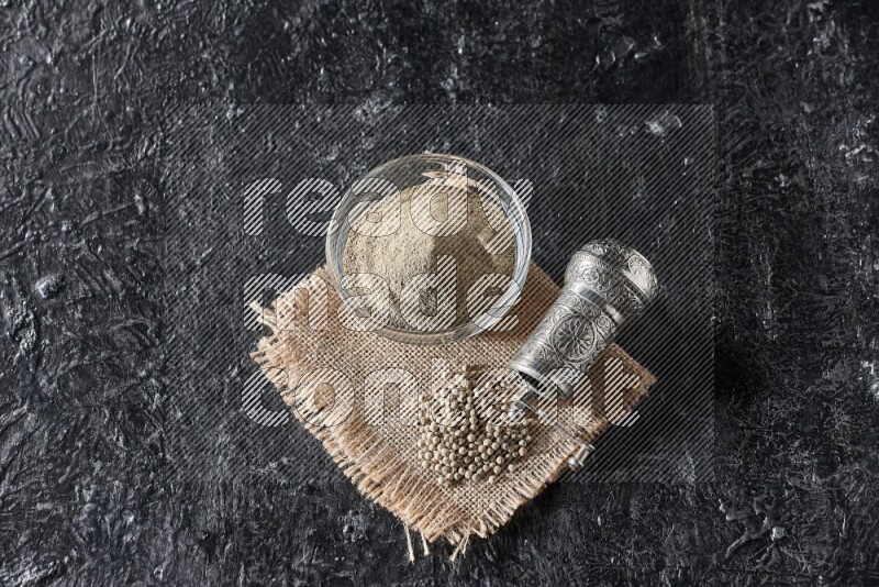 A glass bowl full of white pepper powder with white pepper beads on a burlap piece of fabric and a metal grinder on textured black flooring