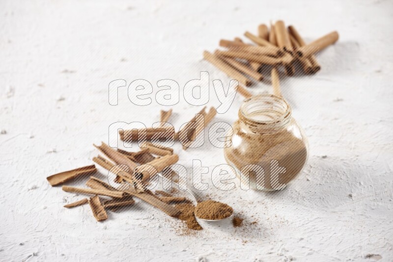Herbal glass jar full cinnamon powder and a metal spoon surrounded by cinnamon sticks on a white background