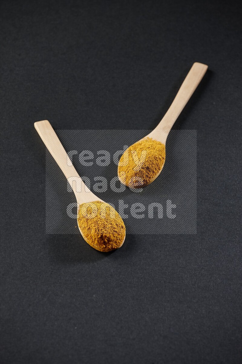 2 wooden spoons full of turmeric powder on black flooring