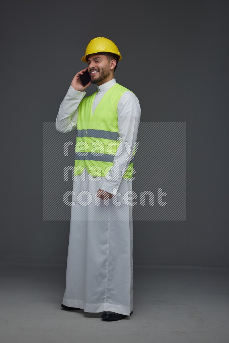 A Saudi man wearing Thobe with a yellow safety vest and white helmet standing and talking in the phone eye level on a gray background