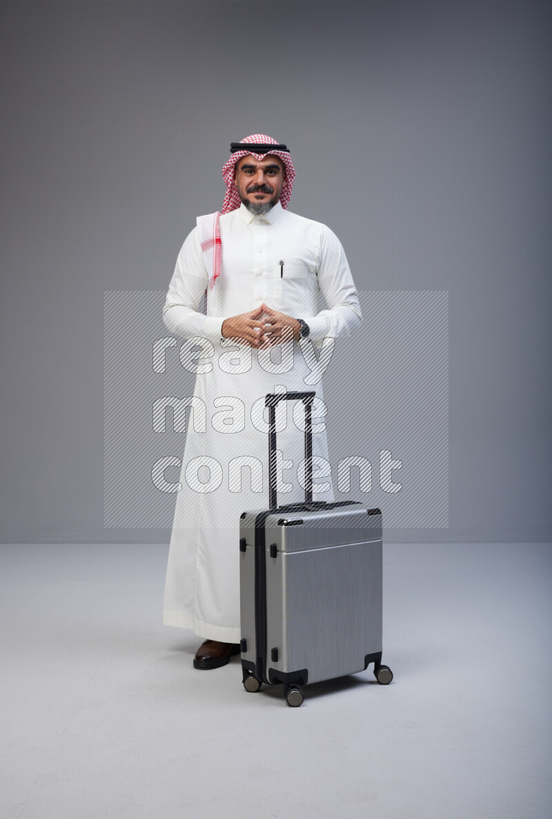 Saudi man wearing Thob and red Shomag standing holding Travel bag on Gray background