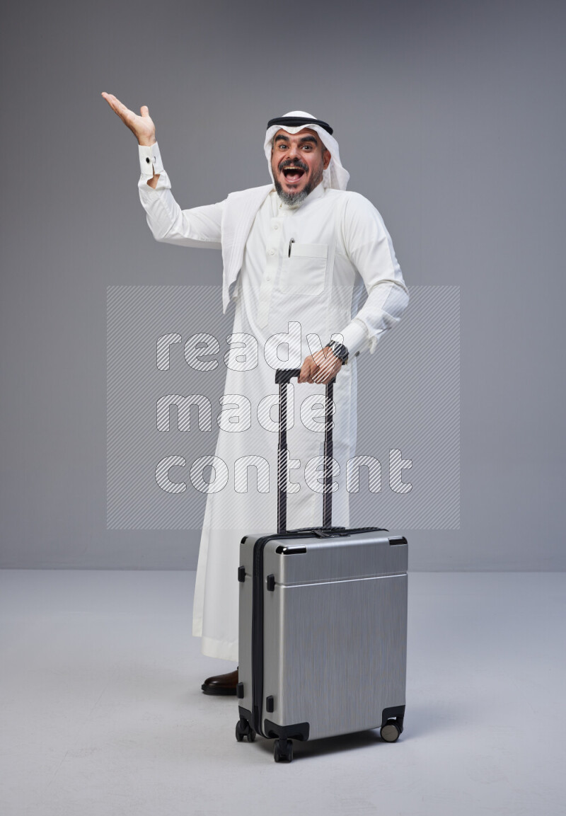 Saudi man wearing Thob and white Shomag standing holding Travel bag on Gray background