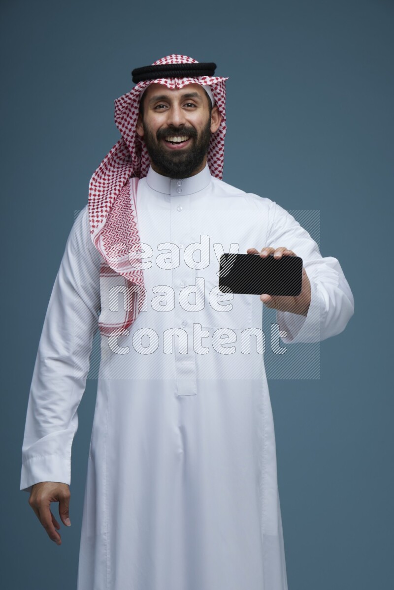 A man showing his phone Screen in a blue background wearing Saudi Thob with Shomag