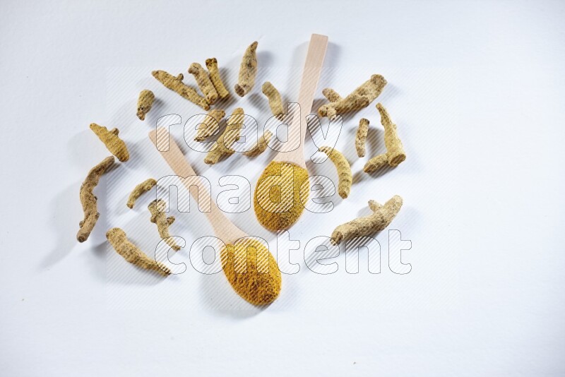 ملعقتان خشبيتان ممتلئتان ببودرة الكركم مع أصابع الكركم المجففة بجانبهما على خلفية بيضاء