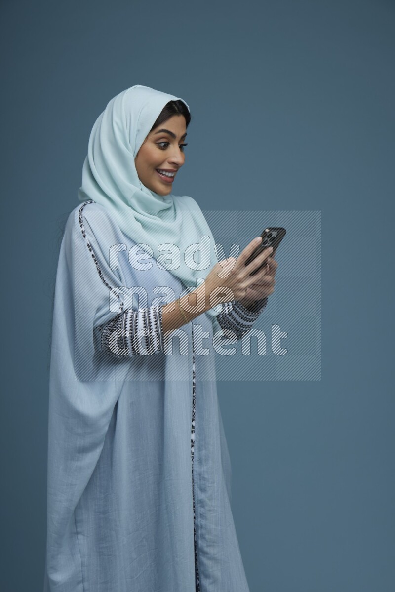 A Saudi woman Texting in a blue background wearing a blue Abaya with hijab