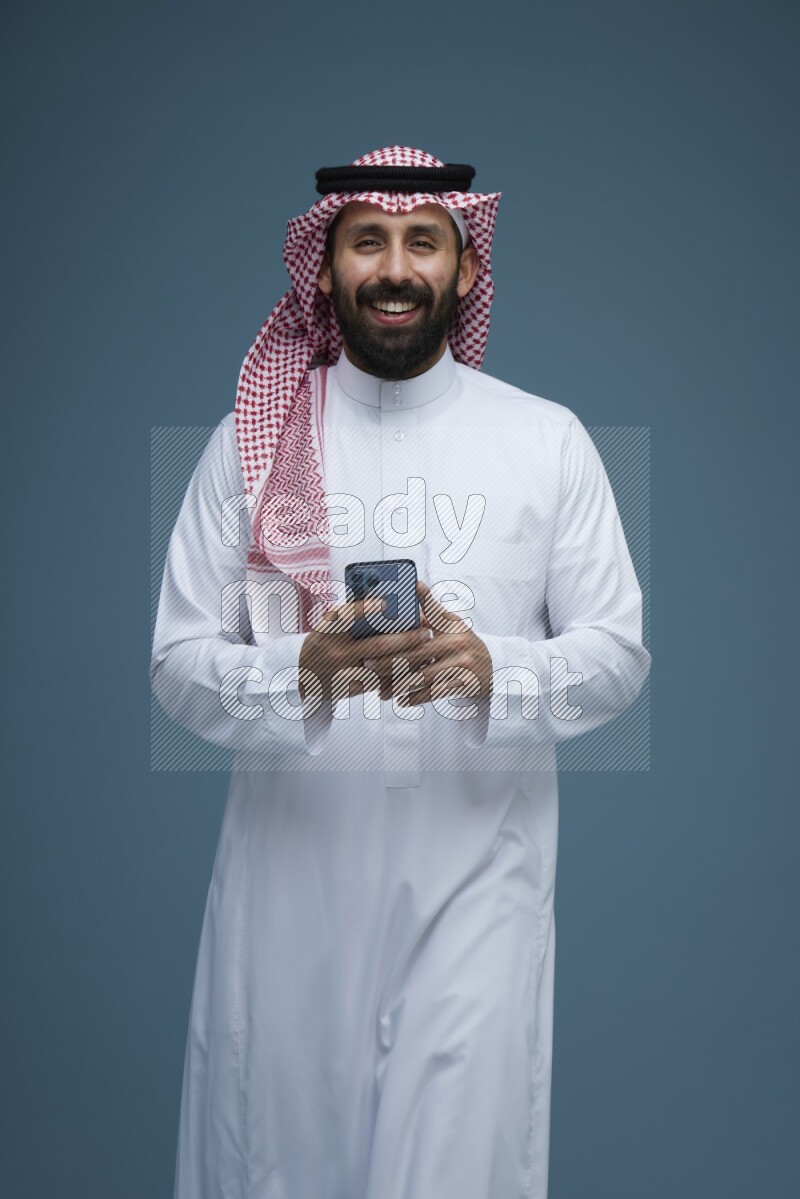 A Male posing with a phone in a blue background wearing Saudi Thob and Shomag