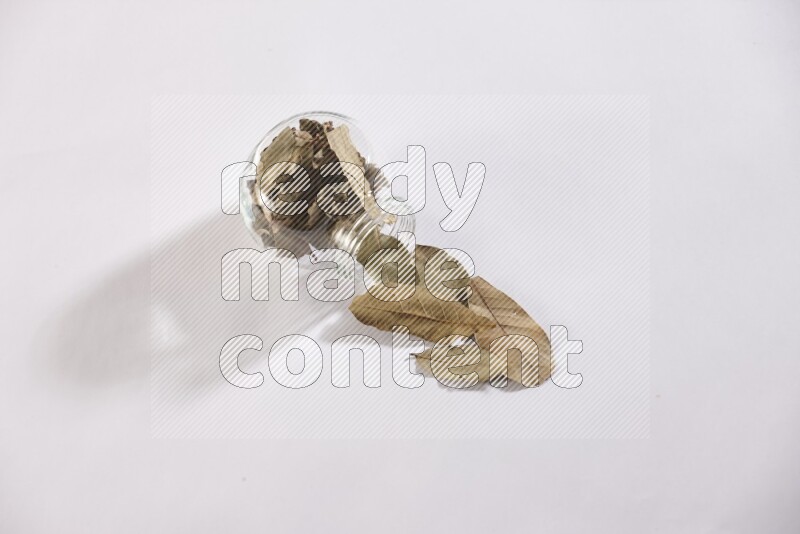 A glass spice jar full of dried bay leaves and jar is flipped and leaves spread out on white flooring