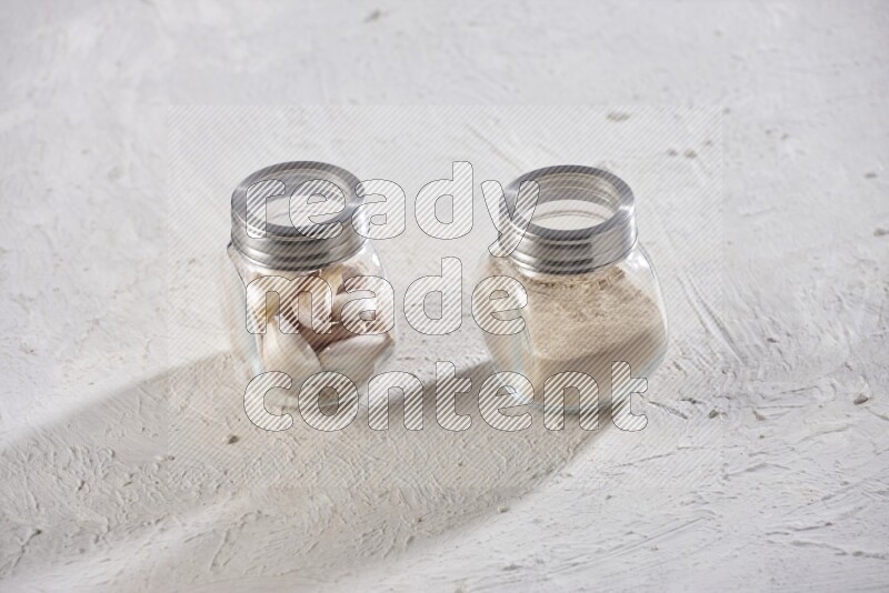 2 glass spice jars one full of garlic powder and the other full of cloves on a textured white flooring